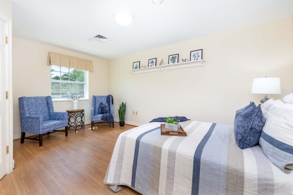 Spacious resident bedroom at Artis Senior Living of Branford with two blue upholstered armchairs, a window seating nook, bed with striped bedding, and coastal wall art shelf