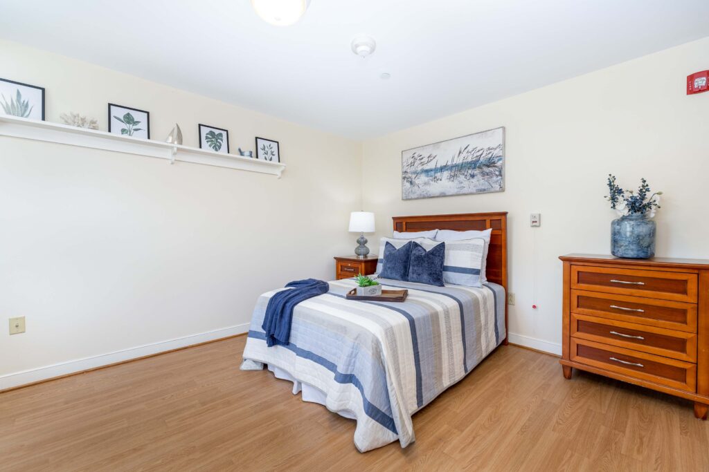 Resident bedroom at Artis Senior Living of Branford with wood bed frame, blue and white striped bedding, dresser, nightstand, and coastal-themed wall art and shelf decor