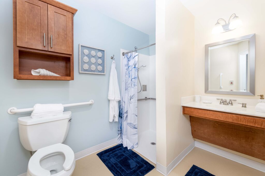 Accessible resident bathroom at Artis Senior Living of Branford with grab bars, walk-in shower, wall-mounted vanity, and coordinated blue and wood decor