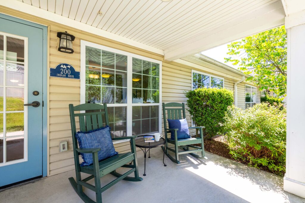 Covered porch outside the Indian Neck neighborhood entrance at Artis Senior Living of Branford, with two green rocking chairs, blue throw pillows, and a small side table