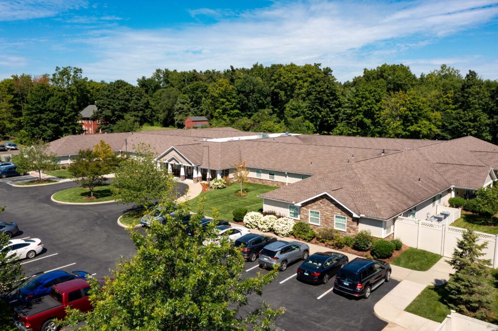 Aerial view of Artis Senior Living of Branford memory care community showing side entrance, resident parking lot, and stone-accented single-story building bordered by trees