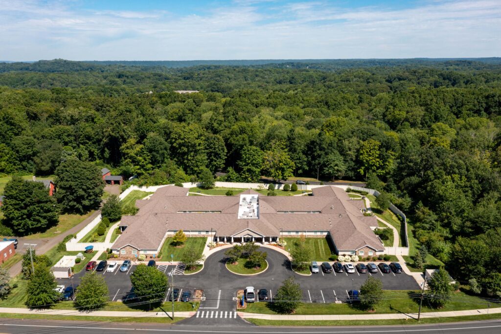 Aerial view of Artis Senior Living of Branford memory care community surrounded by wooded landscape, showing front entrance, circular driveway, secure fenced courtyard, and parking areas