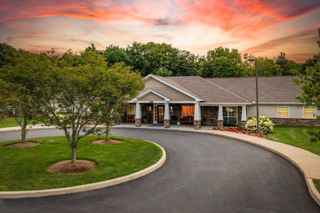 Single-story senior living community entrance with stone accents, white columns, covered front porch, and curved driveway at sunset — Artis Senior Living of Branford memory care community